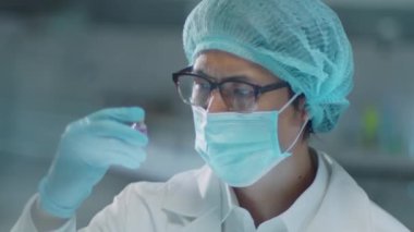 Korunaklı yüz maskesi, tıbbi şapka ve eldivenli Asyalı erkek kimyagerin laboratuarda çalışırken mor kimyasalları incelerkenki göğüs görüntüsü.