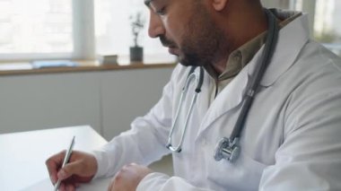 Tıp belgelerini dolduran Orta Doğulu erkek doktor, modern ofisin beyaz masasında oturuyormuş gibi eğiliyor.