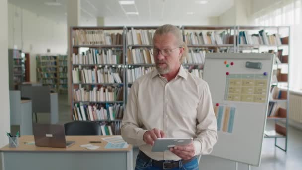 デジタルタブレットを使った中年の白人男性教師のミディアムショット 図書館の学生グループに英語の文法を説明 — ストック動画