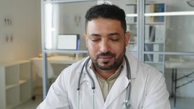 Orta Doğulu orta yaşlı, beyaz önlüklü, kameraya gülümseyen, modern klinikteki ofisinde masasında oturan beyaz erkek doktorun yavaş yavaş portresi.