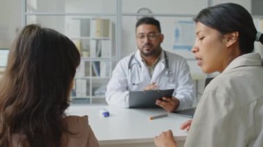 İlgili bir anne, kızının alnını ısıya göre kontrol ediyor ve modern klinikteki sağlık randevusu sırasında Orta Doğulu erkek çocuk doktoruyla konuşuyor.