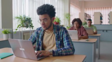 Dişli gömlek ve gözlük takan, dizüstü bilgisayarda çalışan ve kütüphanedeki diğer uluslararası öğrencilerle ders alırken öğretmenlere soru sormak için elini kaldıran genç çift ırklı adamın beline dikkat edin.