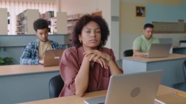Modern kütüphanede dizüstü bilgisayar başında oturan ve kameraya bakan, çok ırklı sınıf arkadaşlarıyla birlikte İngilizce öğrenen genç Afrikalı Amerikalı kız öğrencinin belden yukarı portresi.