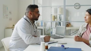 Orta yaşlı Orta Doğulu erkek çocuk doktorunun belden yukarı bakışı. Doktor muayenehanesinde küçük kızı olan İspanyol bir kadınla konuşuyor.