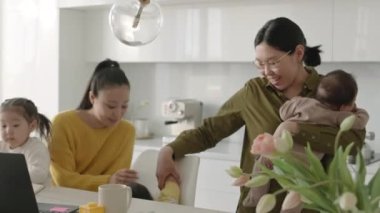 Kadın arkadaşı ve çocuklarıyla evde otururken kucağında bebeği sallayan orta boy bir Asyalı kadın resmi.