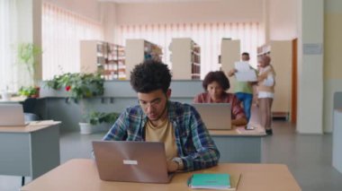 Çift ırklı ve Afrika kökenli Amerikalı erkek ve kız öğrencilerin kütüphanede sıralarında dizüstü bilgisayarlarla otururken sınıf arkadaşlarının bulanık arka planda sohbet etmesi.