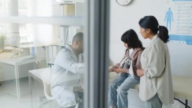 Orta boy İspanyol küçük bir kız elinde yumuşak oyuncakla tıbbi kanepede otururken pediatri muayenehanesinde muayeneden geçerken ve annesinin yanında durup doktoru dinlerken.