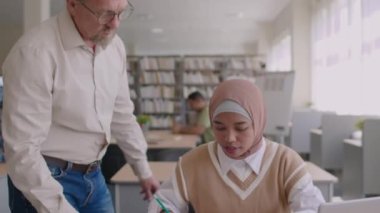Yetişkin beyaz erkek öğretmen, kütüphane dersi sırasında Müslüman kadın göçmen öğrencinin İngilizce dil bilgisi ödevi yapmasına yardım ediyor.