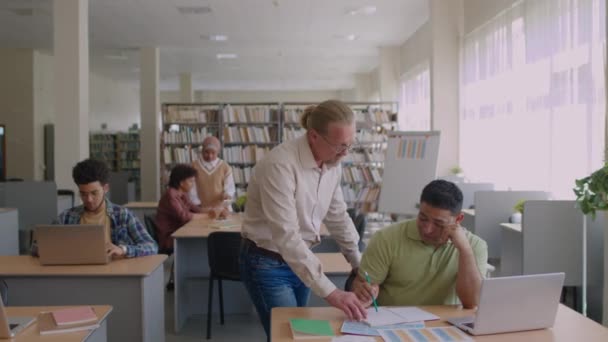 現代の図書館でのグループレッスン中に中東からの移民学生が文法ワークシートを行うのを支援する 高齢の男性英語教師の中間ショット — ストック動画