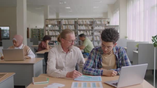 大人の白人男性のチューターに耳を傾けるBiracial移民の学生のミディアムショット現代図書館で彼に英語の文法を説明 — ストック動画