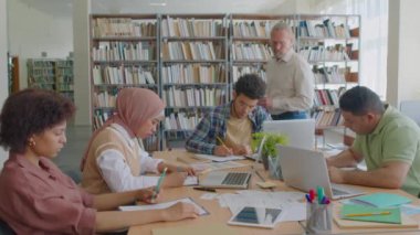 Modern kütüphanede birlikte oturup test yapan bir grup göçmen öğrencinin orta halli fotoğrafı. Bu arada olgun beyaz erkek öğretmen onları izliyor ve izliyor.
