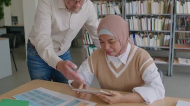 Orta boy, tesettürlü Müslüman kız öğrencinin sınıfta dijital tablet üzerinde egzersiz yaparken olgun beyaz erkek öğretmenin ona yardım ettiği bir fotoğraf.