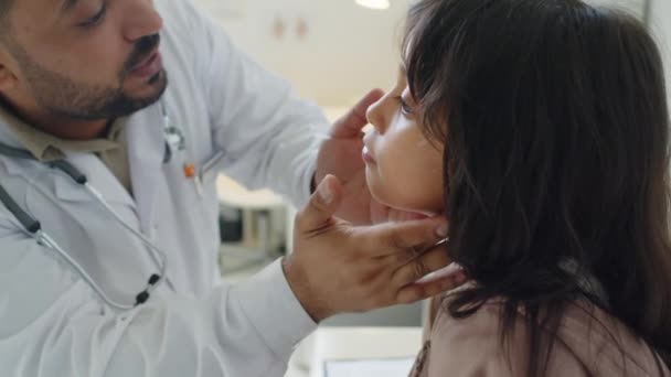 中东男性耳鼻喉科医生用手电筒检查在诊所预约检查的拉美裔小女孩甲状腺和喉咙时的中度特写 — 图库视频影像