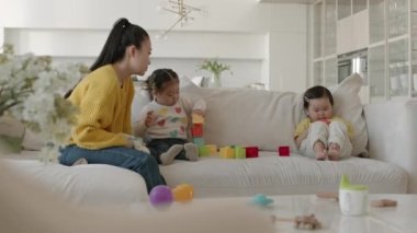 Modern oturma odasındaki beyaz kanepede yan yana otururken renkli yapı taşları olan iki küçük kızıyla oynayan mutlu Asyalı kadının orta boy fotoğrafı.