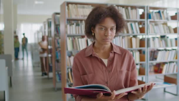 アフリカ系アメリカ人女性大学生の中央スローモの肖像画は 鮮やかな図書館の背景に立ってカメラで笑顔で手の厚い百科事典を持っています — ストック動画