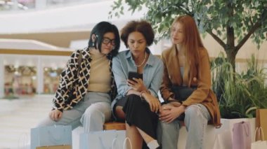 Çağdaş alışveriş merkezindeki bankta oturmuş akıllı telefonlara bakarak sohbet eden çok ırklı üç genç arkadaşın orta boy fotoğrafı.