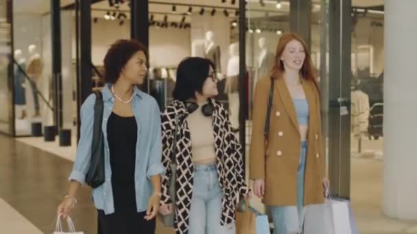ショッピングバッグを携帯する現代的なショッピングセンターを歩いている間 3人の若い多人種間のガールフレンドのミディアムショット — ストック動画