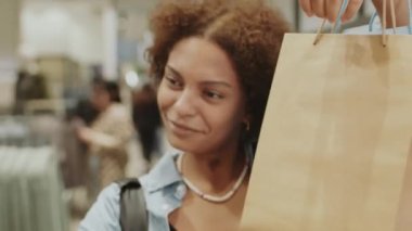 Şık giysi dükkanında arkadaşlarıyla görüntülü sohbet ederken akıllı telefon kamerasında alışveriş torbaları sergileyen mutlu genç siyahi kadının belini kaldırın.
