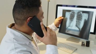 Orta ölçekli Orta Doğulu erkek radyoloji uzmanı meslektaşıyla telefonda konuşurken modern tıp ofisinde çalışan hastaların akciğer röntgeni sonuçlarını tartışıyor.