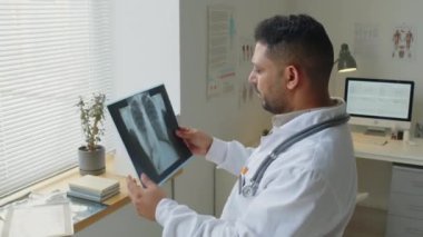Orta yaşlı Orta Doğulu beyaz önlüklü erkek doktorun hastanın akciğer röntgenini incelerken orta boy fotoğrafı.