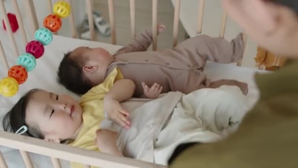 アジアの女性の肩越しに 彼女の2人の赤ん坊の子供を昼寝のためにベビーベッドに入れた ベビーベッドの赤ん坊の兄の隣に横たわっている間 カラフルな掛かるラトルのおもちゃで遊ぶ小さな女の子 — ストック動画