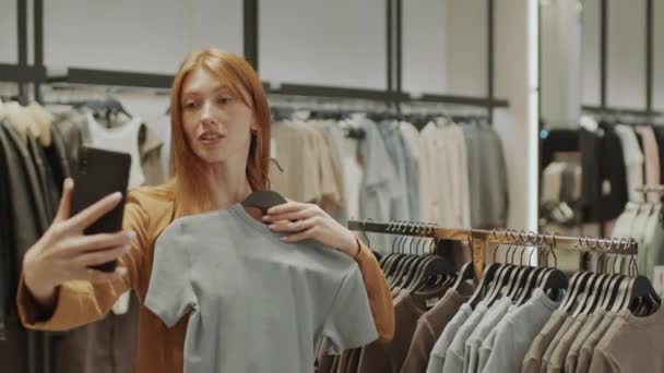 若い白人ジンジャーハイレッド女性のミディアムショット 服店でTシャツを選択し スマートフォンでビデオチャットを介して友人に助言を求める — ストック動画