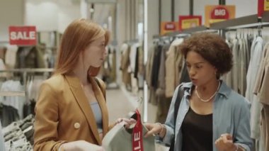 Mevsimsel indirimler sırasında kıyafet mağazasında alışveriş yaparken iki çok ırklı kadının giysi giyip tartıştığı orta boy bir fotoğraf.