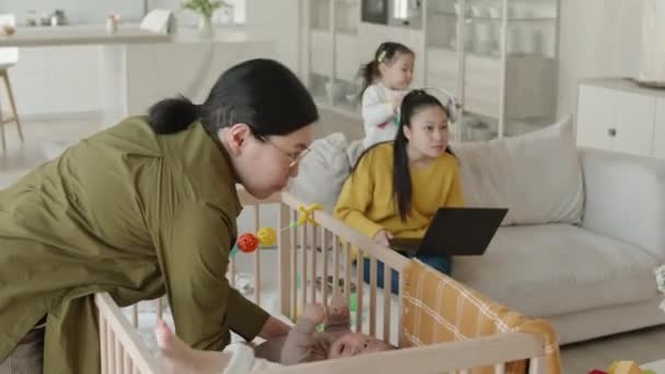 自宅で赤ん坊を抱えたアジア人女性2人の中型ショット 赤ちゃんの息子をベビーベッドで眠らせようとしている一方 2番目は 遊び心のある小さな娘がヘッドフォンで気を散らそうとしているラップトップで作業しようとします — ストック動画