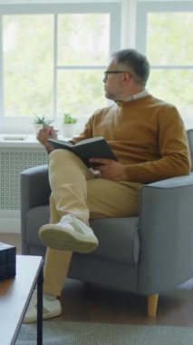 Ofisindeki koltukta oturan deneyimli erkek psikoloğun dikey görüntüsü. Pencereden bakıyor ve deftere notlar yazıyor.