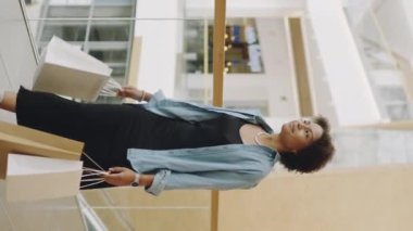 Kendine güvenen Afrikalı Amerikalı genç bir kadının dikey yavaşlığı elinde bir sürü kese kağıdıyla çağdaş alışveriş mağazasında yürüyor.