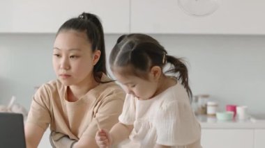 Minimalist beyaz modern mutfakta annesinin yanında oturan küçük kızı defterine çizim yaparken Asyalı genç bir kadının dizüstü bilgisayarla çalışmasını belden yukarısını görmek.