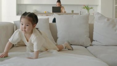 En sevdiği yumuşak oyuncak tavşanıyla ilgilenen küçük Asyalı kızın evde oynarken battaniyeyle nazikçe örttüğü orta boy bir fotoğrafı. Annesi arka planda dizüstü bilgisayarda çalışıyor.