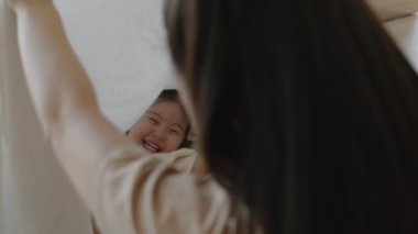 Neşeli bir Asyalı anne ve komik küçük kızının sırtında battaniyeyle oynuyorlar, sıcak evde birlikte vakit geçiriyorlar.