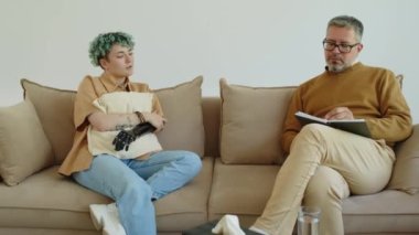 Protez kollu Z kızı yastığa sarılıyor ve terapi sırasında koltukta otururken kıdemli psikoloğun sorularını cevaplıyor.