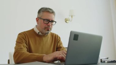 Ofisinde çalışırken laptopta yazan gri saçlı olgun erkek psikoloğun orta boy fotoğrafı.