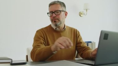 Orta yaşlı, gri saçlı, şık giyinen, gözlüklü, dizüstü bilgisayarlı ve ofisteki telefonlara cevap veren bir adamın orta yaşlı fotoğrafı.