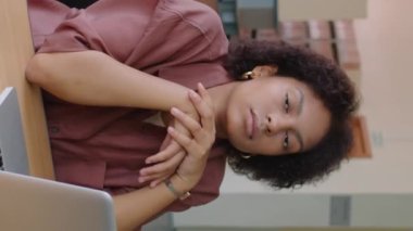 Modern kütüphanede dizüstü bilgisayarın yanında oturan ve kameraya bakan genç Afrikalı Amerikalı kız öğrencinin dikey bel üstü portresi.