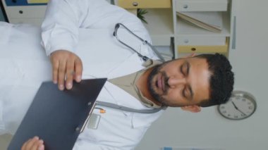 Medikal ofiste elinde panoyla kameraya poz veren kendinden emin Orta Doğulu erkek çocuk doktorunun dikey yavaş portresi.