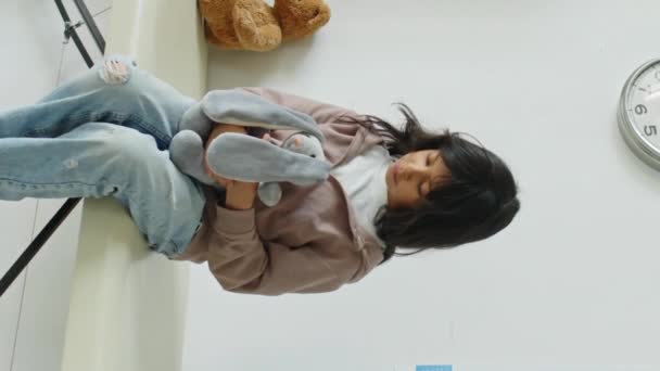 軽い医者のオフィスの医学のソファに座って 任命を待っている手の柔らかいおもちゃを持つ勇敢なヒスパニック少女の垂直媒体の遅い肖像画 — ストック動画