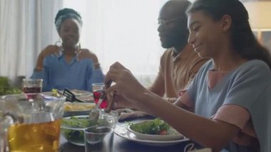Afro-Amerikalı kız evde ailesiyle yemek yerken tabağa salata koyup gülümsüyor.