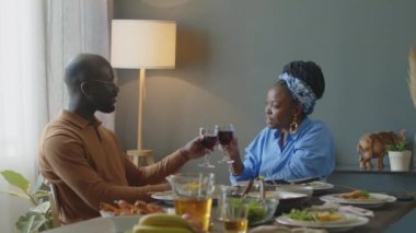 Mutlu Afro-Amerikan aile çifti kadeh tokuşturuyor, kırmızı şarap içiyor ve evde şenlikli bir akşam yemeğinde sohbet ediyorlar.