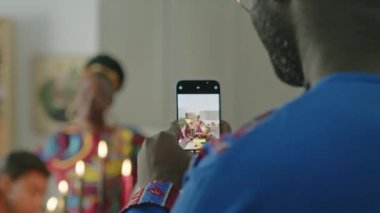 Afrika kökenli Amerikalı bir adamın Kwanzaa kutlamalarında aile telefonuyla fotoğraf çekerken, neşeli bir eş poz verirken ve dans ederken kızlar Kinara mumlarına bakarken.