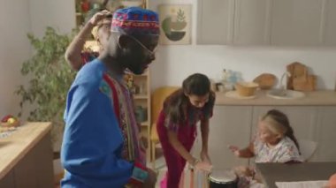 Kwanza bayramı kutlamaları sırasında oturma odasında dans eden neşeli aileleri Dashiki kıyafetleri içinde davul çalan iki küçük Afrikalı Amerikalı kız.