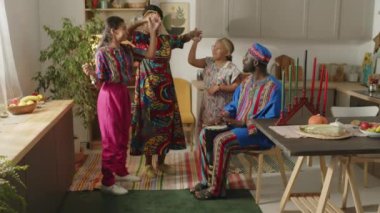 Afrika kökenli Amerikalı bir babanın Dashiki kıyafetleri içinde ve kufi gülüşü ve davul çalarken mutlu bir eş ve iki küçük kızın Kwanzaa 'daki evinde dans edişinin tam bir yavaş çekimi.
