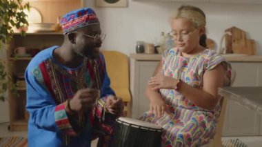 Küçük Afrikalı Amerikalı bir kızın Djembe davul çalmayı öğrenirken babasıyla Dashiki kaftan ve Kwanzaa tatilinde kufi şapkası taktığı orta boy bir fotoğraf.