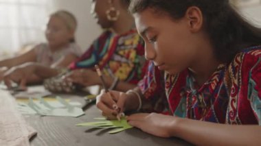 Siyah kızın annesinin ve kız kardeşinin yanında oturduğu ve Kwanzaa kutlamaları için el yapımı tebrik kartına dilek tuttuğu seçmeli bir fotoğraf.