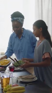 Afro-Amerikan anne ve iki kızın evde aile yemeği hazırlarken yemek masasına ev yapımı yemek koydukları dikey bir fotoğraf.