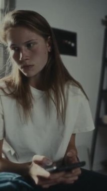 Vertical shot of pissed off teenage girl sitting in bedroom, reading text messages, feeling nervous and throwing phone away