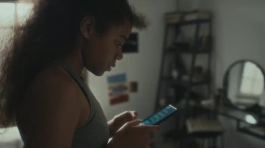 Sinirli Afrikalı Amerikalı genç kız cep telefonuyla mesajlaşıyor ve evde yatak odasında endişeleniyor.