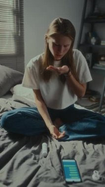 Bir avuç dolusu hap alan, akıllı telefonun yanında uzanan ve siber zorbalık mağduru olarak ağlayan depresif kızın dikey görüntüsü.